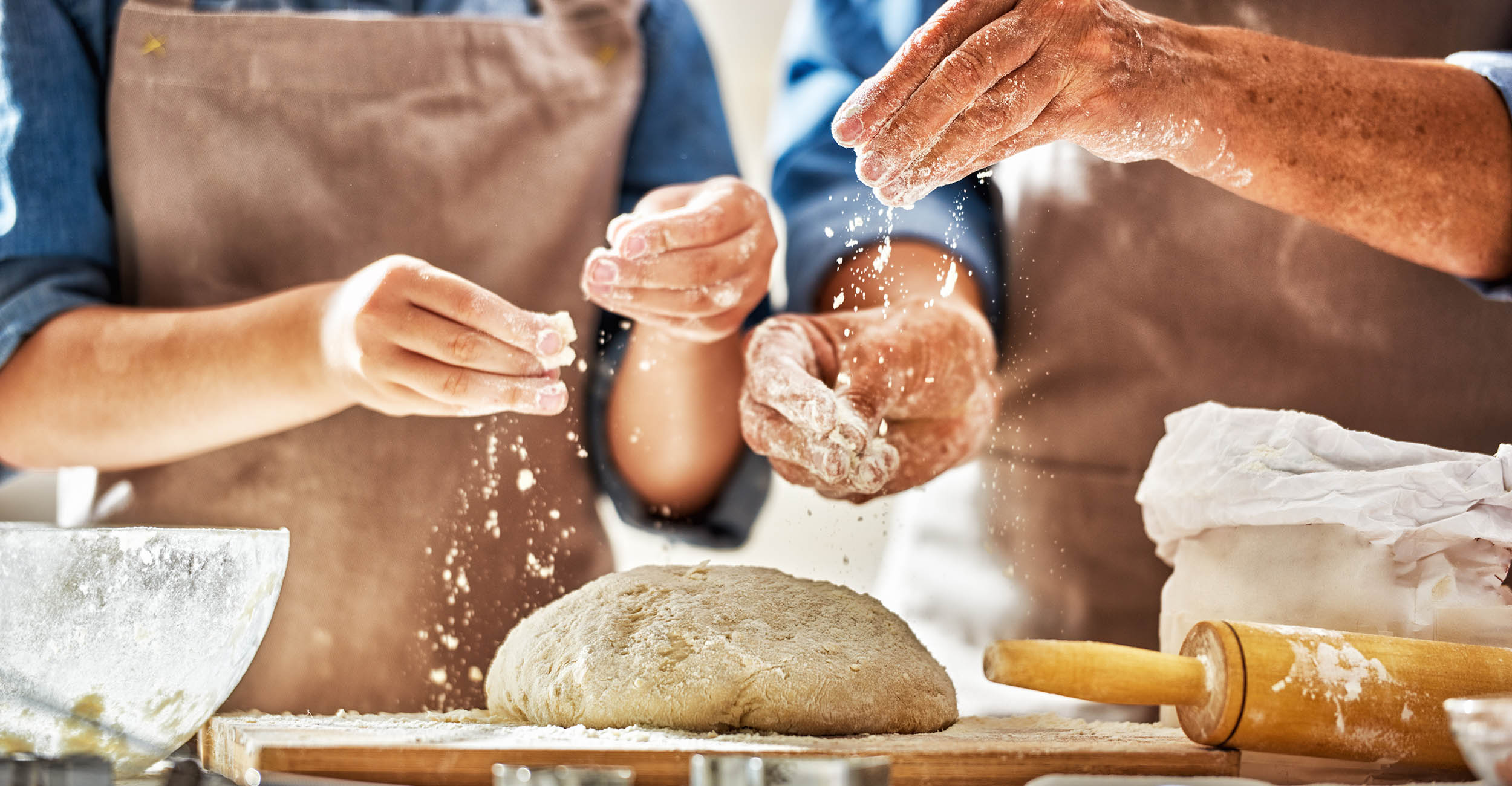 Hands sprinkling flour on bread dough on a kitchen counter with a rolling pin laying nearby