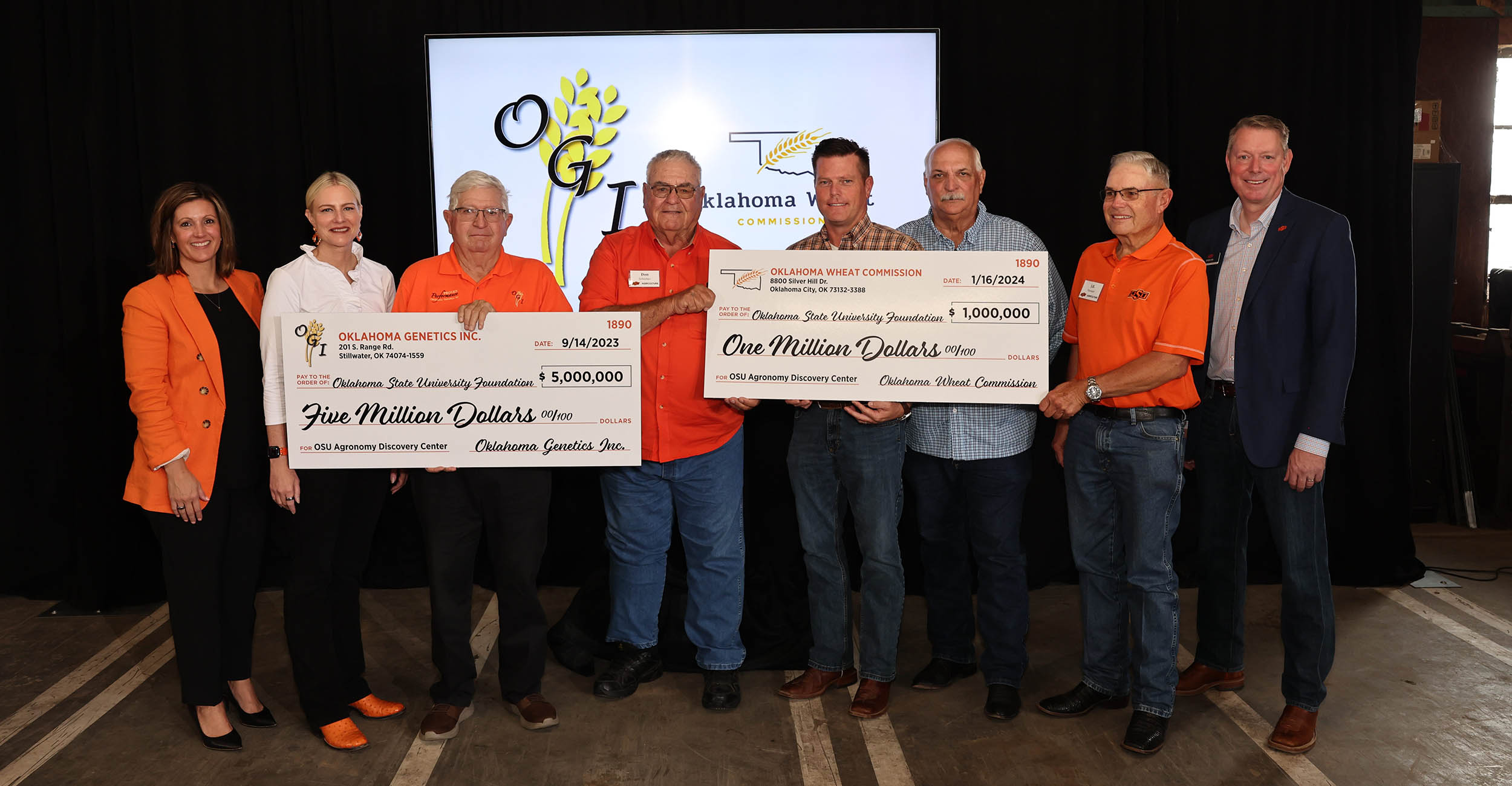 A group of six men and two women wearing orange, white or black shirts stand together holding two large photo prop checks in front of a black backdrop with a screen that displays the Oklahoma Genetics Inc. and Oklahoma Wheat Commission logos.