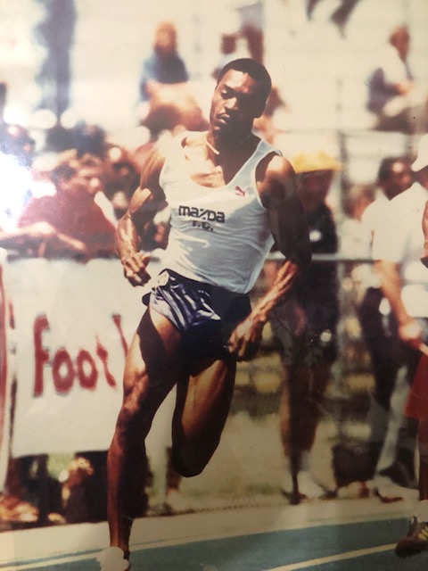 James Butler runs in a sprint on a track. He's wearing blue shorts, a white tank top and track shoes. James Butler runs in a sprint on a track. He's wearing blue shorts, a white tank top and track shoes.